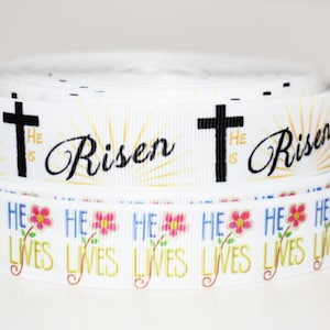 May include: White ribbon with a repeating pattern of a cross and the words "He is Risen" in black text. A second white ribbon with a repeating pattern of pink flowers and the words "He Lives" in yellow text.