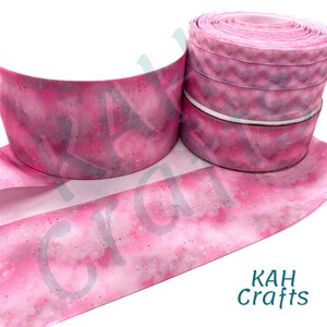May include: Pink and white glitter ribbon with a galaxy print. The ribbon is rolled up and the print is visible on the outside of the roll.