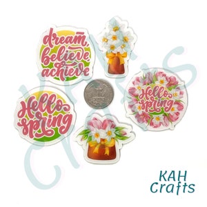 May include: Four colorful stickers with spring-themed designs. One sticker features a bouquet of pink and white flowers in a brown pot with the text "Hello Spring". Another sticker features a bouquet of white flowers in a brown pot with the text "dream, believe, achieve". The remaining two stickers feature the text "Hello Spring" in a floral wreath.