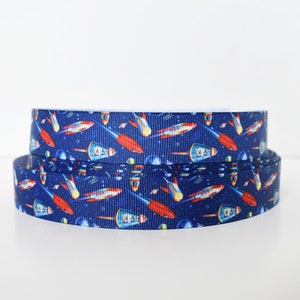 May include: A roll of blue and white ribbon with a repeating pattern of red and white rockets and stars. The ribbon is perfect for adding a space-themed touch to crafts, gifts, and decorations.