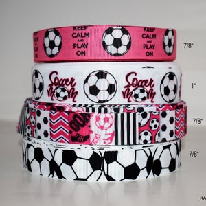 May include: A stack of four pink and white soccer-themed ribbon rolls. The top roll has the text "Keep Calm and Play On". The second roll has the text "Soccer Mom". The third roll has a pink and black geometric pattern. The bottom roll has a white background with black soccer balls. The rolls are 7/8 inch and 1 inch wide.