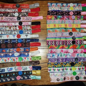 May include: A collection of colorful fabric face mask extenders with buttons. The extenders feature various patterns and designs, including floral, animal print, and tie-dye. Some extenders have text such as "Nurses make it all better!" and "Nursing is a work of heart!"