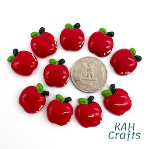 May include: Ten red apple-shaped resin cabochons with green stems and leaves. The cabochons are approximately the size of a US quarter.