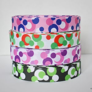 May include: Four rolls of patterned ribbon stacked on top of each other. The ribbons feature a repeating pattern of colorful circles in pink, white, green, purple, and black. The ribbons are likely used for crafting or gift wrapping.