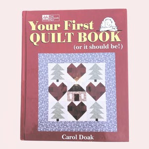May include: A maroon book titled "Your First Quilt Book (or it should be!)" features a quilt design with hearts, trees, and a house. The author's name, Carol Doak, is printed at the bottom. The book has a floral patterned border.
