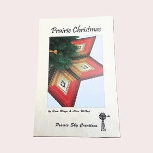 May include: A rectangular instructional booklet titled Prairie Christmas by Pam Weege and Alice Wilhoit. The cover features a photograph of a star-shaped fabric tree skirt in red, green, and gold patterns placed under a green pine tree.