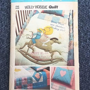 May include: Simplicity pattern 6702 for a Holly Hobbie quilt. The quilt design features a child riding a rocking horse. The pattern includes a pillow and quilt with heart and patchwork designs. The pattern is from American Greetings Corporation.