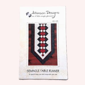 May include: A sewing pattern booklet titled Seminole Table Runner by Atkinson Designs, model SPL-105. The cover features a graphic of a table runner with a geometric pattern in red, black, and white, set against a dark wood background.