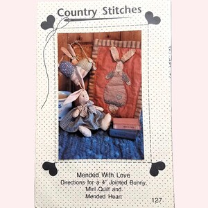 Peut inclure: Un patron Country Stitches avec des motifs de lapin articulé, de mini-couette et de cœur réparé. L'image présente un lapin en peluche dans une robe à carreaux bleus et blancs, et un appliqué de lapin en vichy rouge et blanc sur un oreiller. Le texte dit "Mended With Love."