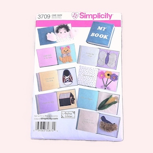 May include: A Simplicity pattern for a fabric book with various illustrated pages. The cover reads "MY BOOK". Inside, pages feature felt appliqués of a dog, shoe, butterfly, and other objects. The pattern is labeled "3709".