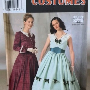 May include: Simplicity Costumes 7312 sewing pattern envelope featuring two dress designs. One is a long-sleeved, dark red plaid dress with a white lace collar. The other is a light blue sleeveless dress with a dark blue sash and small bows.