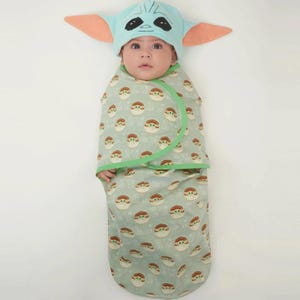May include: A baby wearing a light blue hat with large ears and a matching swaddle blanket. The blanket and hat feature a repeating pattern of a character's face. The blanket is light green with a green trim.