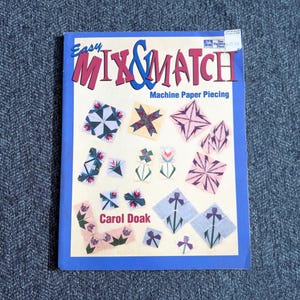 May include: A softcover instructional book titled Easy Mix & Match Machine Paper Piecing by Carol Doak. The cover features a light cream background with a blue border and various colorful geometric quilt block patterns in pink, blue, and green.