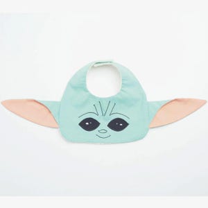 May include: A baby bib in the shape of a character's face, featuring a light teal front with black eyes and a smiling mouth. The bib has attached pink ears on either side, and a white backing.