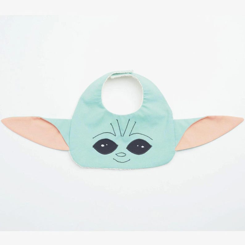 May include: A baby bib in the shape of a character's face, featuring a light teal front with black eyes and a smiling mouth. The bib has attached pink ears on either side, and a white backing.