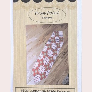 Peut inclure: Un patron imprimé pour un chemin de table saisonnier. Le motif présente un motif géométrique rouge et blanc. Le texte "Prim Point Designs" est en haut et "#400 Seasonal Table Runner" en bas.