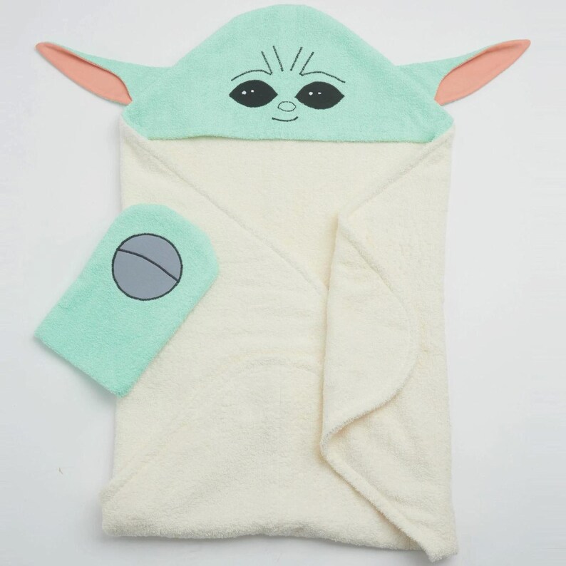 May include: A hooded baby towel and washcloth set. The towel is cream-colored with a light green hood featuring a character's face with large eyes and ears. The washcloth is light green with a gray circle design.