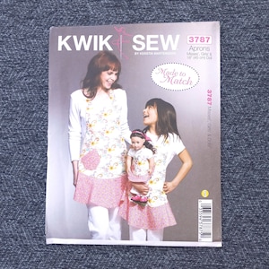 May include: A sewing pattern for aprons, featuring a woman and a girl wearing matching aprons with a floral design and pink trim. The pattern includes sizes for misses, girls, and an 18-inch doll. The cover reads "KWIK SEW 3787".