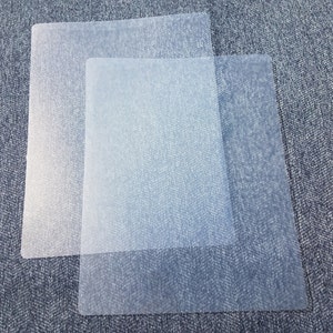 May include: Two translucent, rectangular sheets of plastic are layered on top of each other. The sheets have rounded corners and a slightly textured surface. The color is a pale, almost transparent, blue-gray.