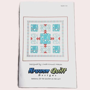 May include: A quilt pattern design on white paper. The quilt design features a central square and surrounding squares in shades of blue and white, with red accents. The text "Kruusn Quilt Designs" is visible.