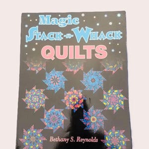 May include: A softcover instructional book titled Magic Stack-n-Whack Quilts by Bethany S. Reynolds. The black cover features a repeating pattern of colorful, star-shaped quilt blocks in shades of blue, pink, and yellow, with white star accents.