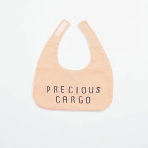 May include: A peach-colored baby bib with the words "PRECIOUS CARGO" printed in black. The bib has a curved shape with two tabs at the top for fastening. The background is plain white.