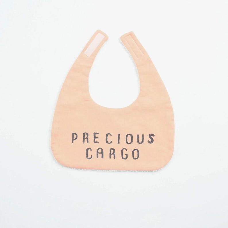 May include: A peach-colored baby bib with the words "PRECIOUS CARGO" printed in black. The bib has a curved shape with two tabs at the top for fastening. The background is plain white.