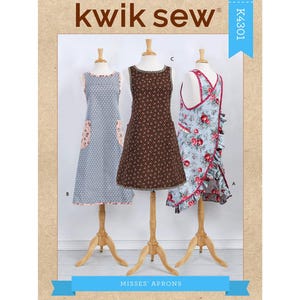 May include: A sewing pattern featuring three different apron designs displayed on wooden mannequins. The aprons include a blue patterned apron with a floral pocket, a brown patterned apron, and a floral apron with red trim. The pattern is labeled "Kwik Sew" and "Misses' Aprons."