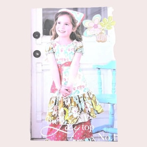 May include: A young girl wearing a floral print dress with a red belt and matching pants. The dress has a ruffled skirt and short sleeves. The outfit is accessorized with a matching headscarf. The text "the Lady top No.1" is visible.
