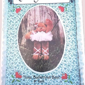 May include: A craft pattern titled "Loving Stitches" features a brown ballerina bear wearing a white tutu and ballet shoes. The bear is 8 inches tall and is set against a wooden background within an oval frame. The frame is surrounded by a blue floral pattern.