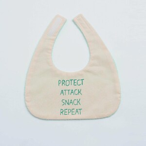 May include: A light pink baby bib with a green trim and the words "PROTECT ATTACK SNACK REPEAT" in green. The bib has a hook and loop closure at the neck.