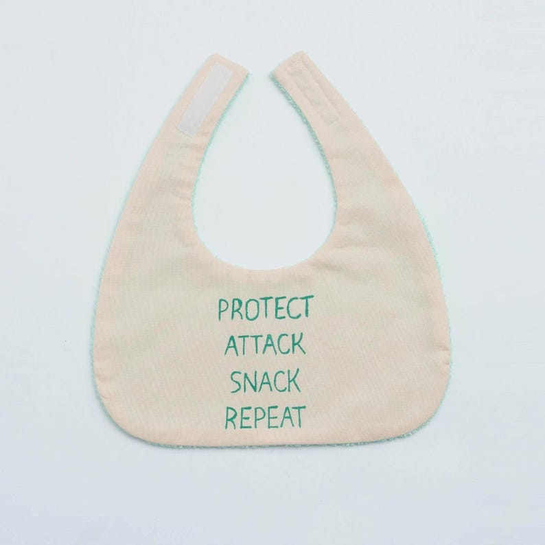 May include: A light pink baby bib with a green trim and the words "PROTECT ATTACK SNACK REPEAT" in green. The bib has a hook and loop closure at the neck.
