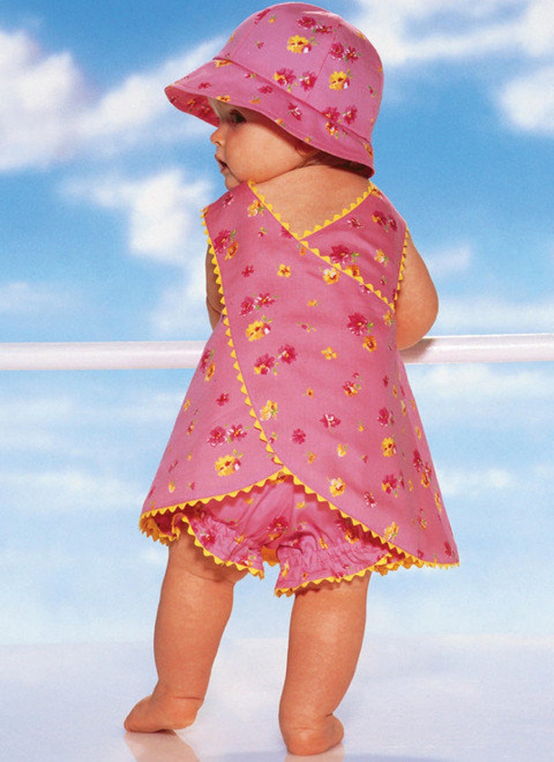 May include: A baby wearing a pink floral print sun hat and matching wrap-style dress with yellow trim. The outfit includes matching bloomers. The baby is standing outside with a blue sky background.