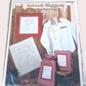 May include: A package featuring a "Redwork Christmas" design by Cindy Taylor Oates. The image shows a white button-down shirt with red trim, a pillow, a drawstring bag, and a quilted square, all with redwork embroidery of Santa Claus.
