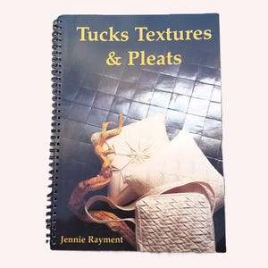 May include: A spiral-bound instructional book titled Tucks Textures & Pleats by Jennie Rayment. The cover features a dark background with images of cream-colored fabric pillows and a small bag, alongside a piece of driftwood.