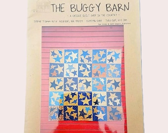 May include: A quilt pattern featuring a grid of squares with star designs in shades of blue, gold, and cream. The quilt is displayed against a red and pink wooden background. The text "THE BUGGY BARN" is at the top.