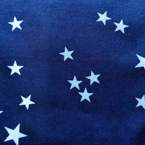 May include: Close-up of a navy blue fabric with a pattern of white stars. The stars are of varying sizes and are scattered across the surface, creating a celestial design. The fabric has a slightly textured appearance.