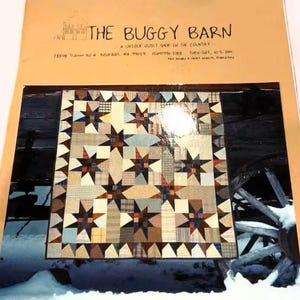 May include: A quilt with a star pattern in shades of brown, blue, and beige. The quilt is displayed on a rustic wooden background with a partial view of a wooden wheel. The text "THE BUGGY BARN" is visible at the top.