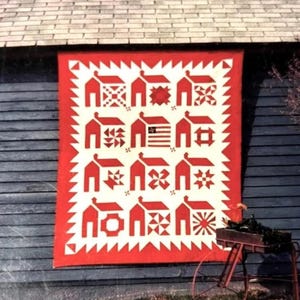 May include: A red and white quilt with a repeating pattern of houses and various geometric shapes. The quilt is hanging on a weathered wooden wall. The quilt's border has a zig-zag design.