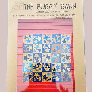 May include: A quilt pattern featuring a grid of squares, each adorned with star designs in shades of blue, gold, and orange. The quilt is displayed against a backdrop of a pink and red wooden surface. The text "THE BUGGY BARN" is visible at the top.