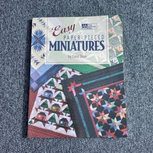 May include: A book titled "Easy Paper-Pieced Miniatures" by Carol Doak. The cover features colorful patchwork quilt designs with geometric patterns, houses, and stars. The book is on a blue textured surface.