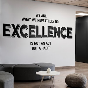 Può includere: Un muro bianco con la citazione "We are what we repeatedly do. Excellence is not an act but a habit." in testo nero. Un divano grigio, un tavolino da caffè bianco e un pouf grigio sono in primo piano.