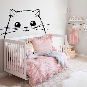 Puede incluir: Una cuna blanca con un juego de cama rosa con volantes y una manta gris. Una pegatina de gato blanco y negro está en la pared sobre la cuna. La palabra "elle" está en un estante sobre la cuna.