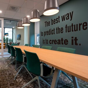 Könnte beinhalten: Ein heller Holzkonferenztisch mit türkisfarbenen Stühlen in einem modernen Büro. Die Wand hinter dem Tisch ist türkisfarben und trägt das Zitat "Der beste Weg, die Zukunft vorherzusagen, ist, sie zu erschaffen." in schwarzen Lettern.