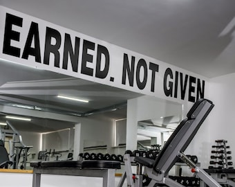 May include: A black and white sign with the text "Earned. Not Given." hanging above a weight bench in a gym. The bench is made of metal and has a black padded seat. The gym is empty and has a white ceiling and walls.