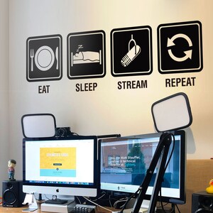 以下が含まれることがあります： 2つのコンピューターモニター、マイク、デスクランプのあるホームオフィスワークスペース。デスクの後ろの壁には、「EAT、SLEEP、STREAM、REPEAT」という黒と白のグラフィックがあります。