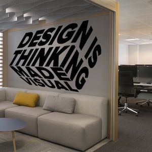 Pode incluir: Um adesivo de parede preto e branco com o texto "DESIGN THINKING IS MADE VISUAL" em estilo 3D. O adesivo está em uma parede cinza em um ambiente de escritório.