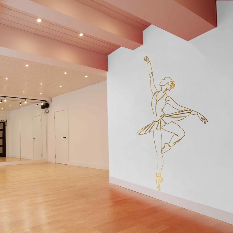 May include: A white wall with a gold outline of a ballerina in a dance pose. The ballerina is wearing a tutu and pointe shoes. The wall is in a room with a wooden floor and a pink ceiling.