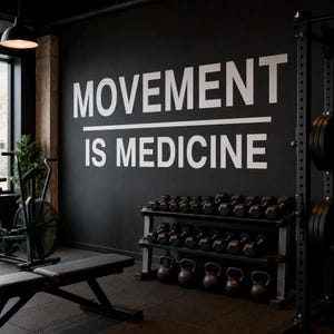 Pode incluir: Uma parede preta com o texto branco MOVEMENT IS MEDICINE e uma linha horizontal entre as palavras. O ginásio inclui uma bicicleta ergométrica preta, um banco de exercícios, um suporte de metal com halteres pretos e kettlebells pretos.