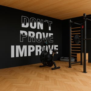 Op de afbeelding: Een sportschool aan huis met een zwarte muur met de motiverende tekst "DON'T PROVE IMPROVE" in het wit. In het midden staat een hometrainer, met een gewichtenrek en houten vloer. Het plafond is van hout.